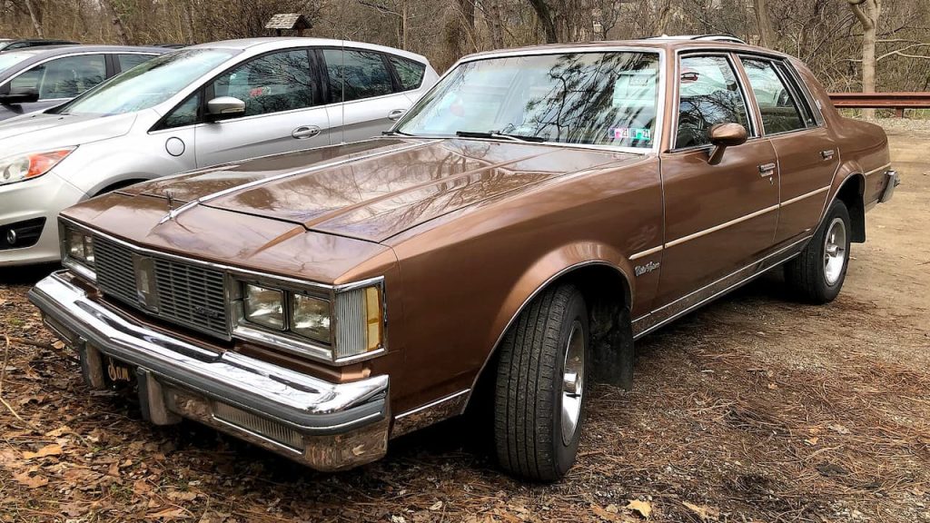 Brown 1985 Oldsmobile Cutlass Supreme sedan with chrome bumpers parked on dirt ground near modern white cars