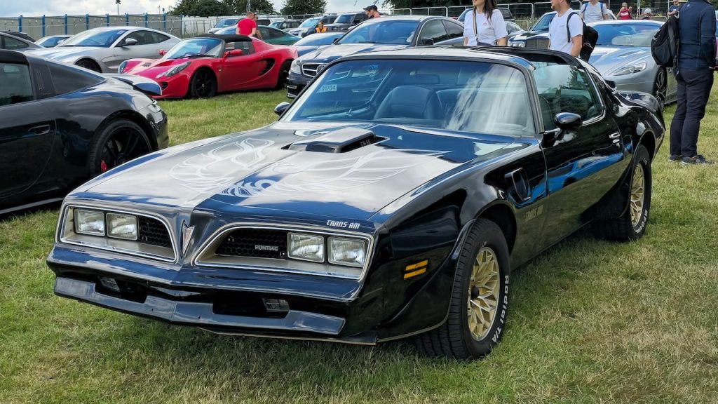 Black Pontiac Firebird Trans Am with hood scoop and gold wheels displayed at outdoor car show among other sports cars