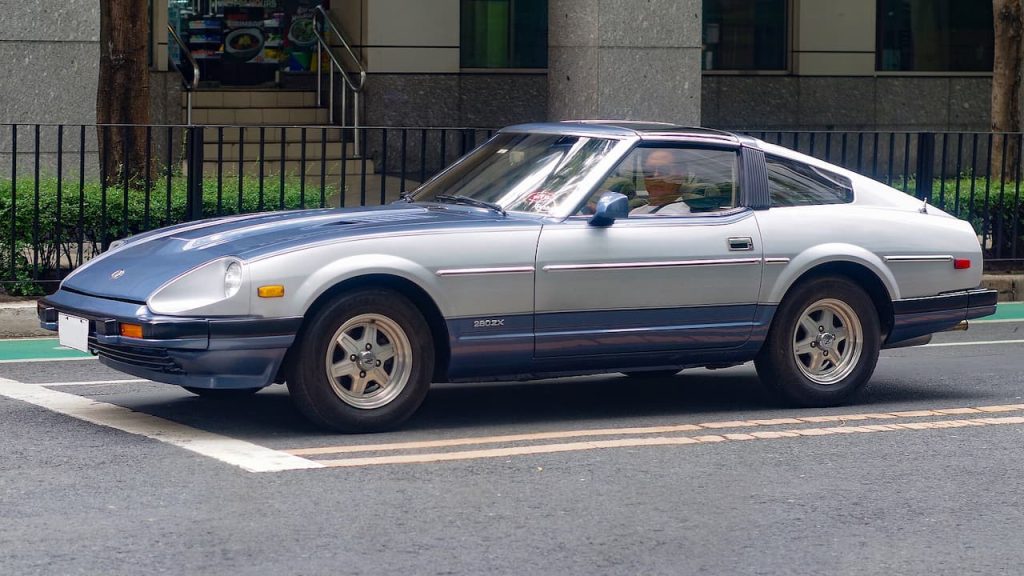 Silver Datsun 280ZX sports car parked on street with apartment building and metal fence in background