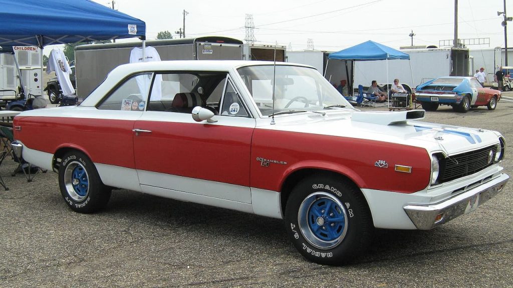Classic red and white 1969 AMC SC/Rambler 390 muscle car with blue racing wheels parked at outdoor car show event