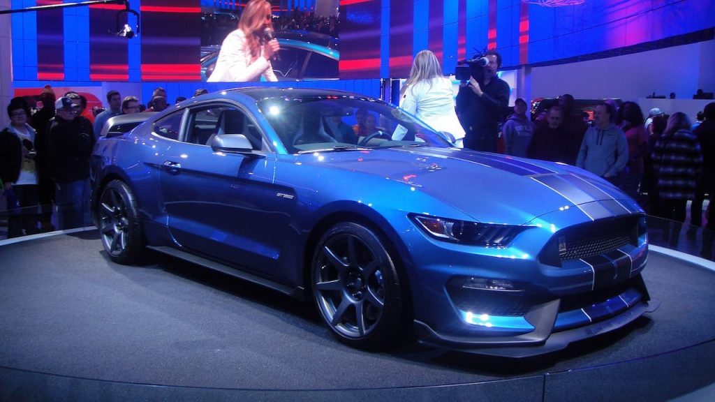 Blue Ford Mustang Shelby GT350R with racing stripes displayed on stage at auto show with audience watching