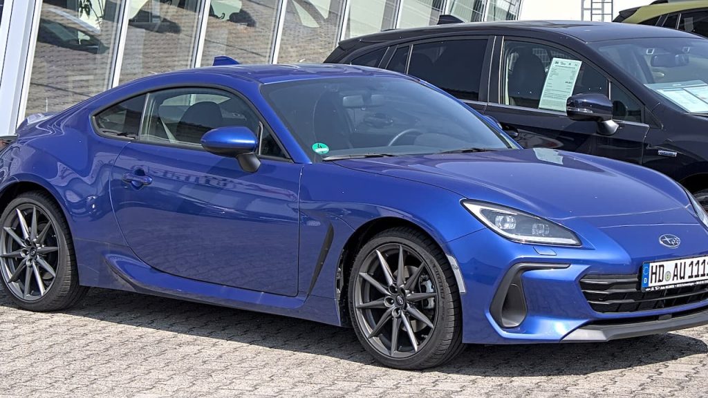 Blue Subaru BRZ sports coupe with alloy wheels parked outside dealership showroom with other vehicles visible in background