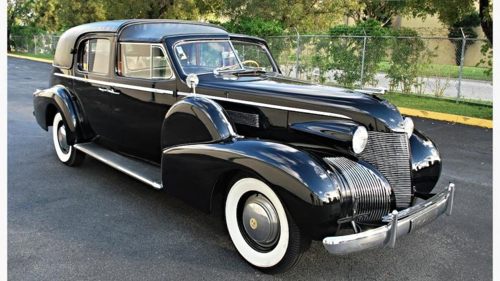 Glossy black 1939 Cadillac Series 75 with whitewall tires, chrome grille and running boards, parked on asphalt near chain-link fence