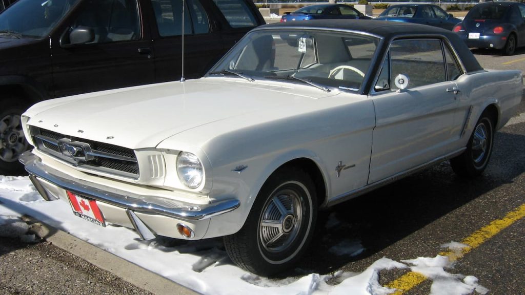 White 1964 Ford Mustang with black roof parked in snowy lot, Canadian license plate visible, modern cars nearby