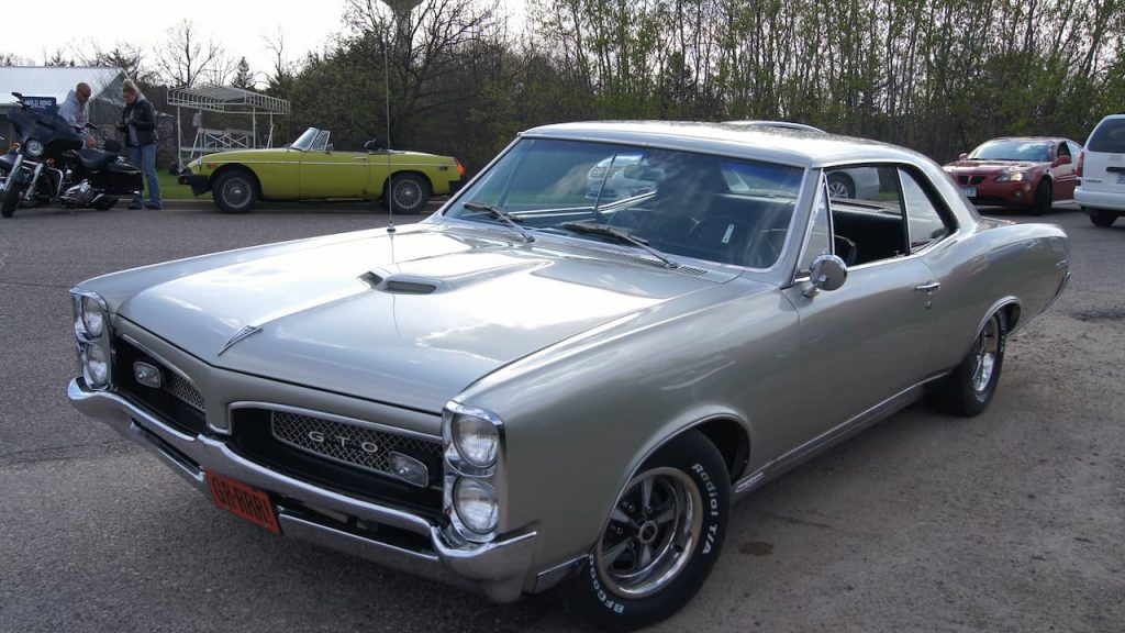 Silver 1967 Pontiac GTO muscle car parked at car meet with yellow convertible and motorcycle visible in background