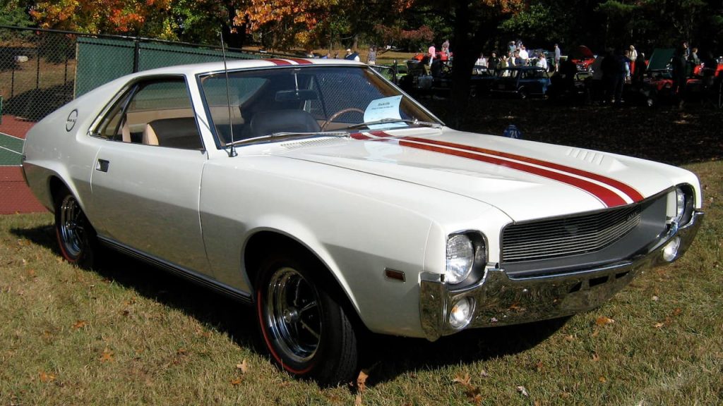White 1968 AMC AMX muscle car with red racing stripes displayed on grass at autumn car show near tennis courts