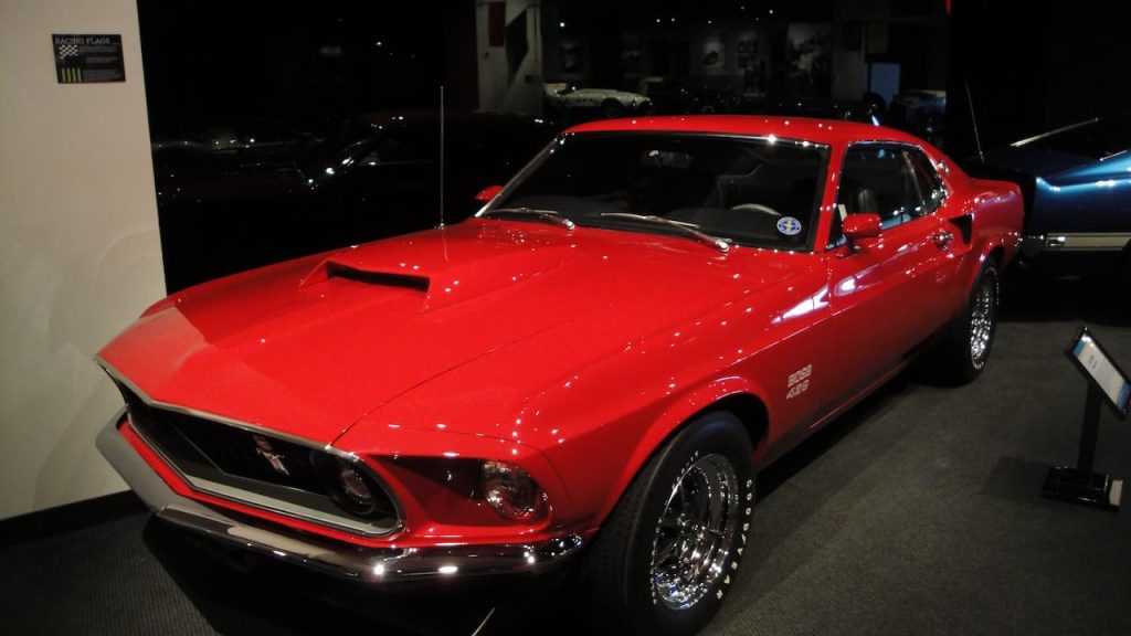 Bright red 1969 Ford Mustang Boss 429 muscle car displayed in dimly lit automotive museum with chrome wheels and hood scoop