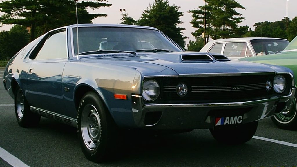 A dark blue 1970 AMC AMX 390 muscle car parked on asphalt with trees and other vehicles in background