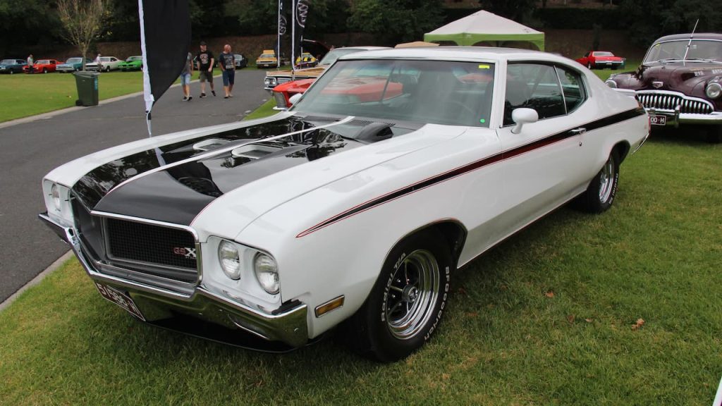 White Buick GSX 455 muscle car with black hood stripe displayed on grass at outdoor car show with spectators nearby