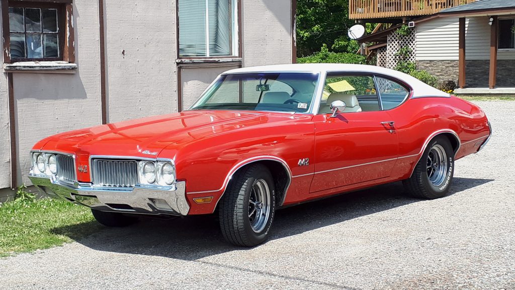 
Red Oldsmobile 442 muscle car with white roof parked in driveway beside white house with wooden deck