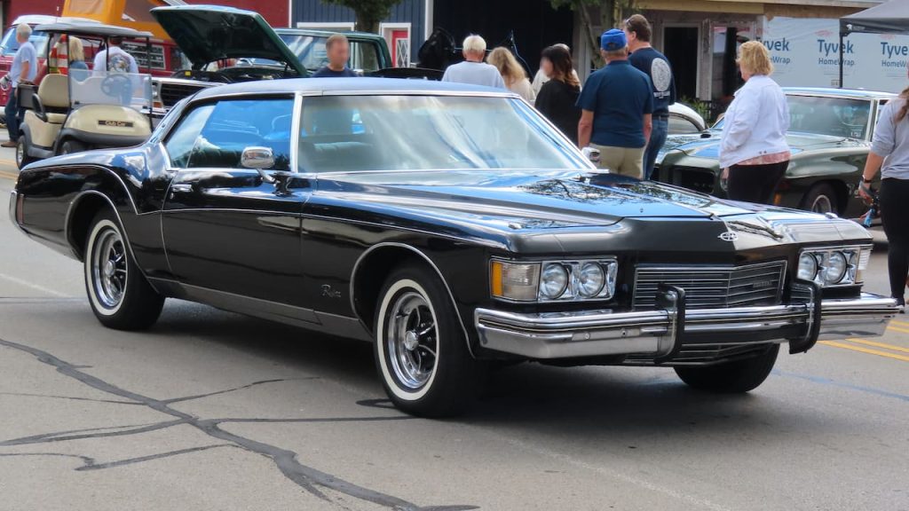 Black 1973 Buick Riviera with chrome trim and white roof displayed at car show with spectators browsing nearby