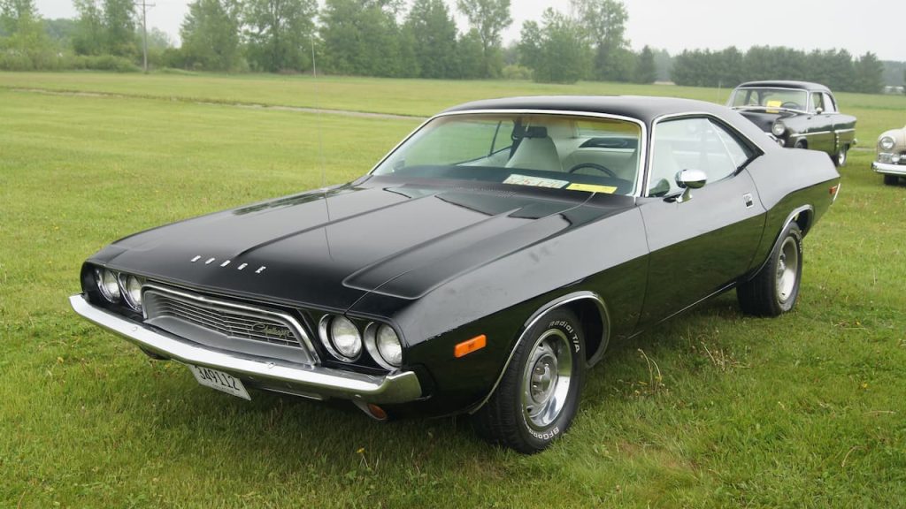 Black 1974 Dodge Challenger muscle car with white racing stripes parked in green field at classic car show event