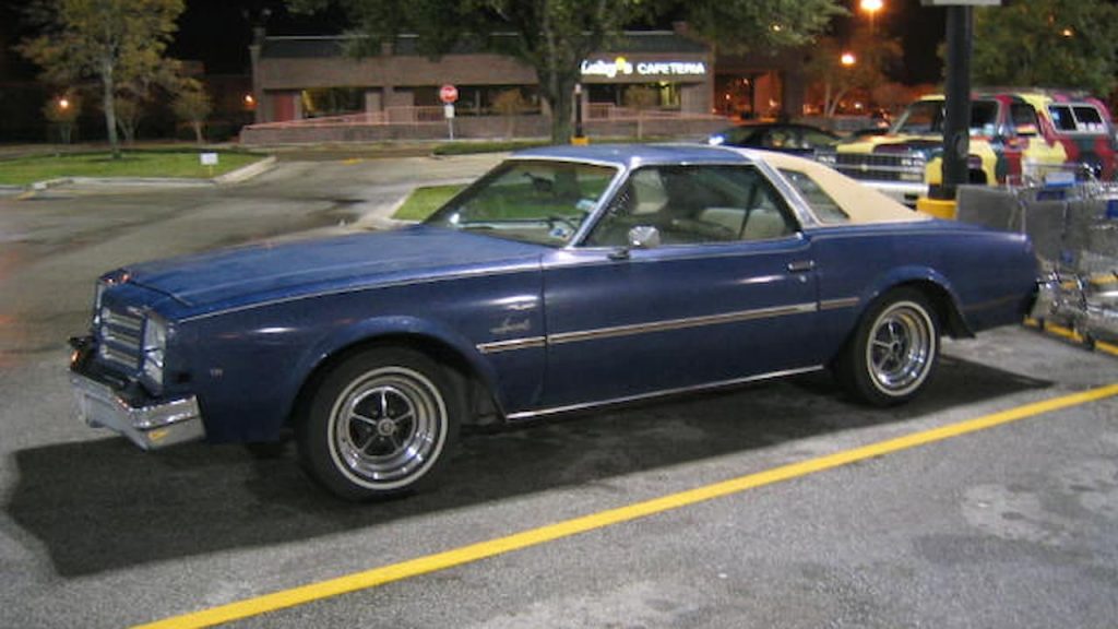 Blue vintage 1977 Buick Century Special 455 muscle car with white convertible top parked in shopping center lot at night under street lights