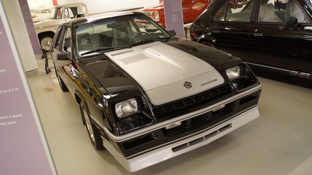 Black and silver 1985 Dodge Charger displayed in automotive museum with other classic cars on white floor