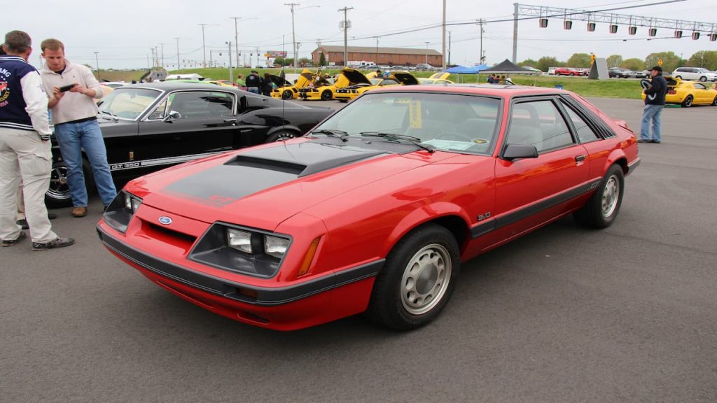 Red 1986 Fox body Mustang with black racing stripes parked at outdoor car show with people and other classic vehicles nearby