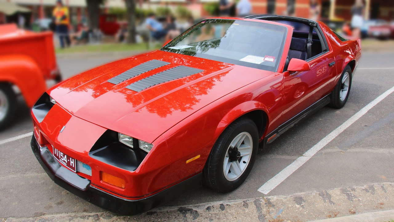 Red 1987 Chevrolet Camaro with gray racing stripes and black wheels displayed at outdoor classic car show
