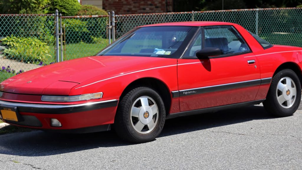 Red Buick Reatta with black roof parked on street beside chain-link fence with green foliage background
