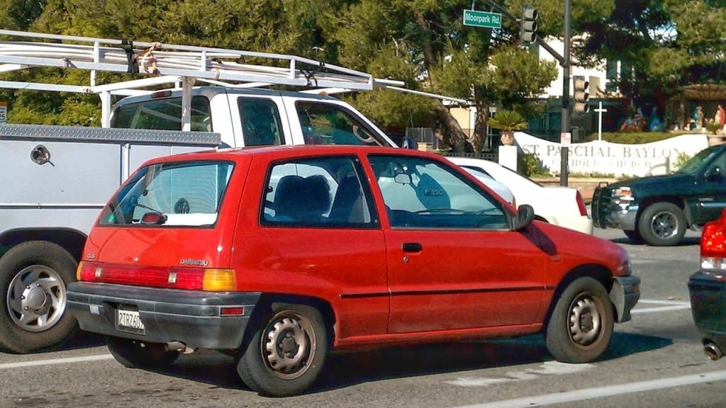 Red 1989 Daihatsu Charade parked on street with white pickup truck and utility vehicle nearby, green street sign visible