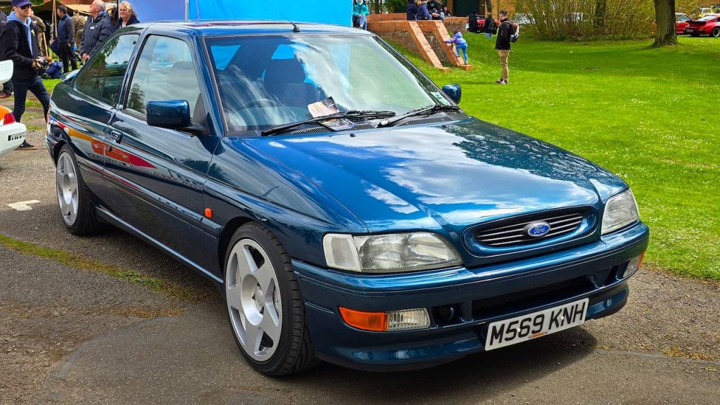 Dark blue 1995 Ford Escort with UK license plate M569KNH parked at outdoor car show with spectators nearby