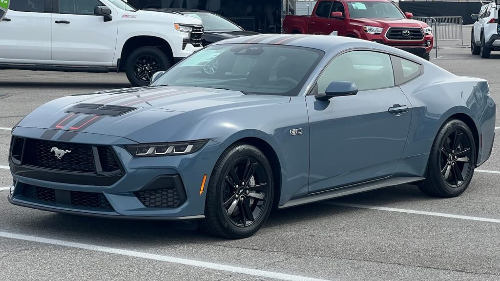 A gray 2025 Ford Mustang GT with black racing stripes parked in a lot beside other vehicles
