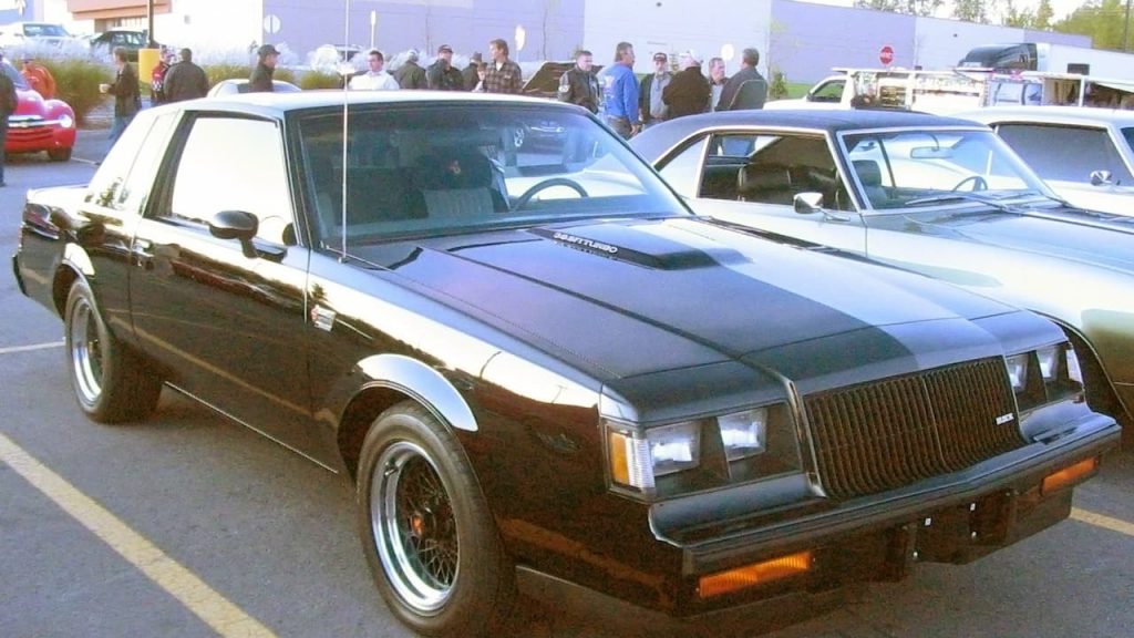 Black 1987 Buick Regal Grand National muscle car with T-top roof at car show with crowd of spectators nearby