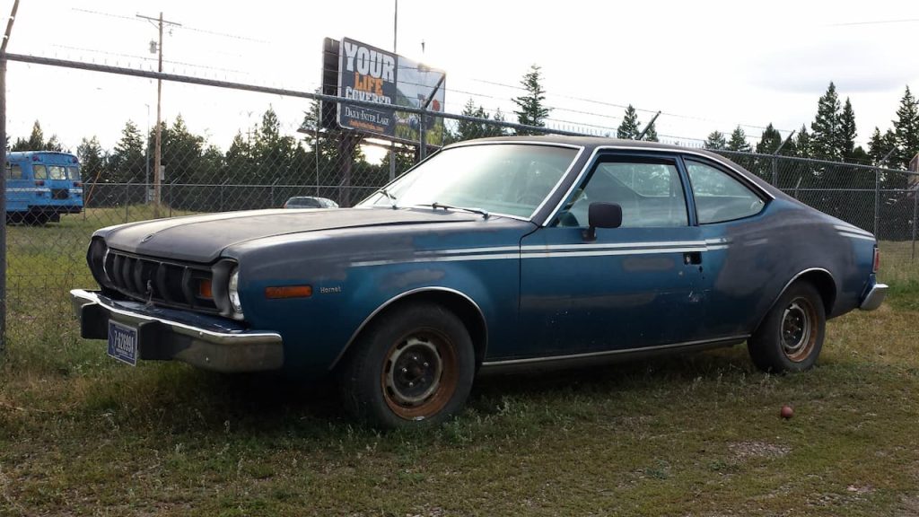 Blue 1984 AMC Hornet parked on grass lot with chain-link fence, billboard, and pine trees in background