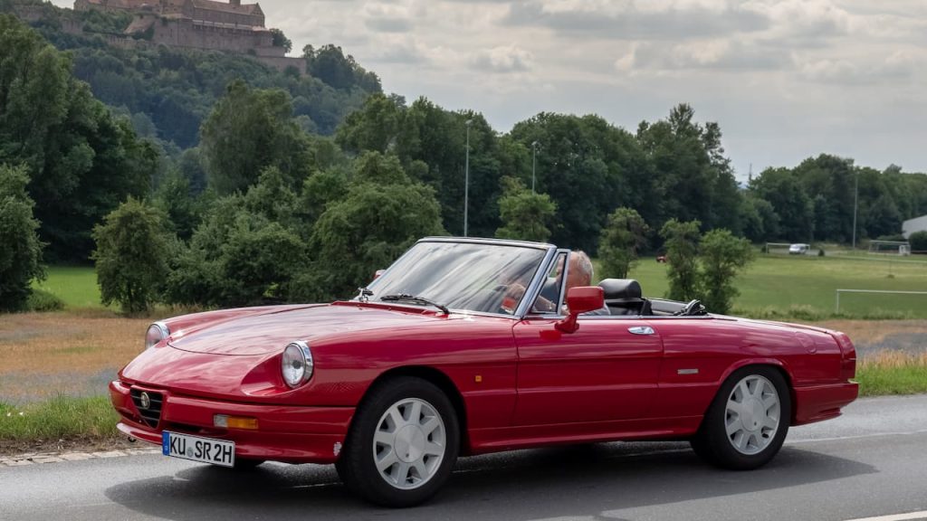 Red Alfa Romeo Spider convertible with registration KU-SR2H parked on roadside with castle, hills, and green countryside in background