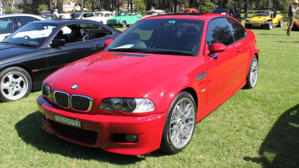 Red BMW M3 E46 coupe with alloy wheels parked on grass at outdoor car show event.