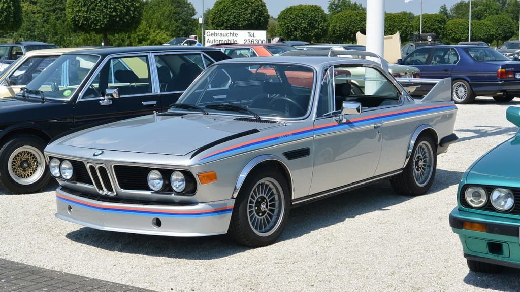Silver BMW E9 CSL with blue and red racing stripes displayed at German used car dealership among other vehicles