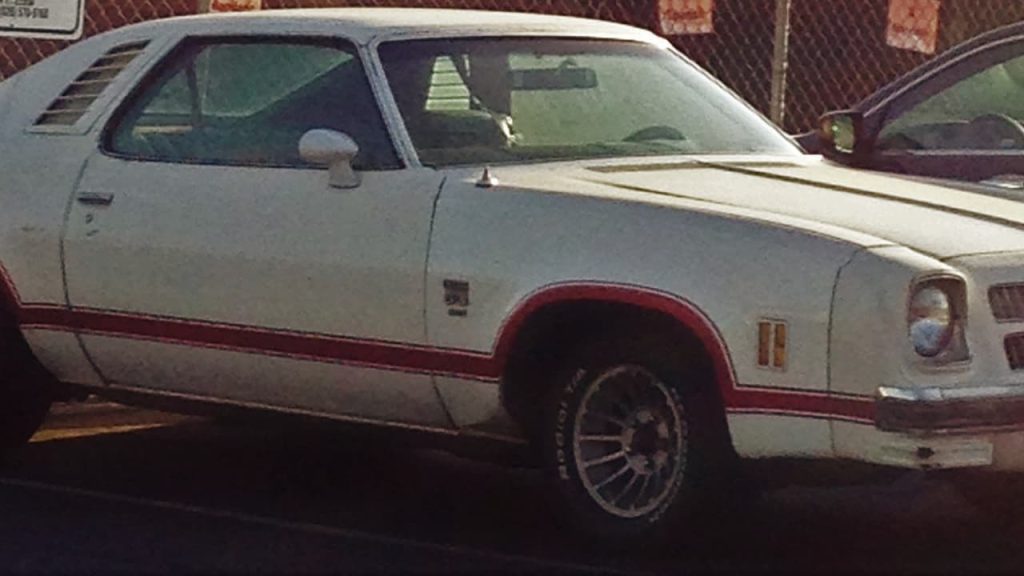 White classic 1976 Chevy Laguna S-3 454 - 245 HP with red trim and side stripes parked outdoors, showing weathered paint and vintage styling