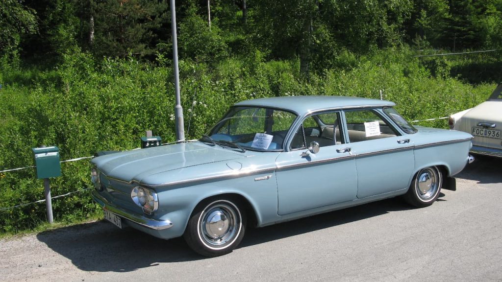 Light blue Chevrolet Corvair four-door sedan with lowered suspension and chrome wheels parked on asphalt near green vegetation