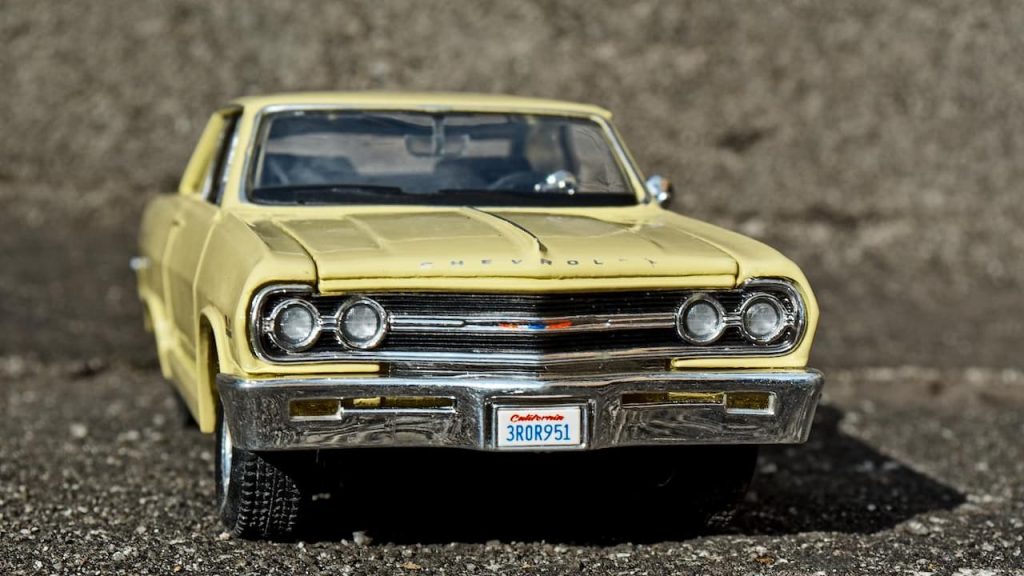 Die-cast model of yellow 1965 Chevrolet Chevelle SS with chrome bumper and California license plate on textured gray surface