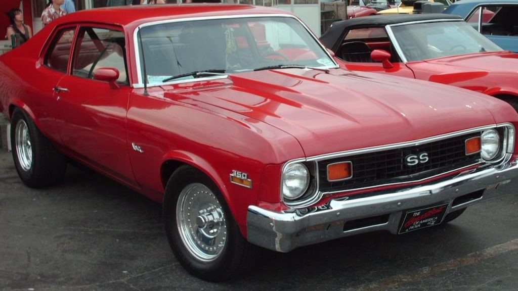 Red Chevrolet Nova with 350 badge and chrome bumper displayed at car show alongside other classic vehicles