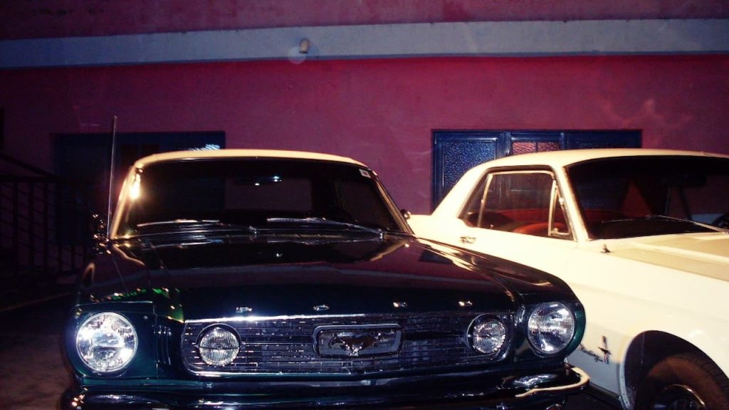 Classic black 1964½ Ford Mustang parked beside white vintage car against burgundy wall with decorative windows at night
