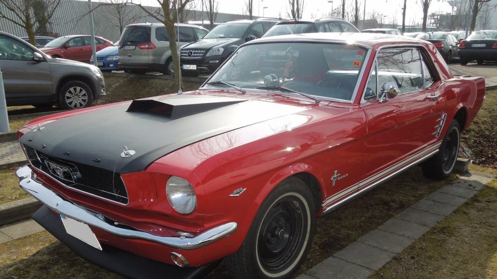 Red Ford Mustang with black hood and racing stripes parked in lot surrounded by modern cars and bare trees