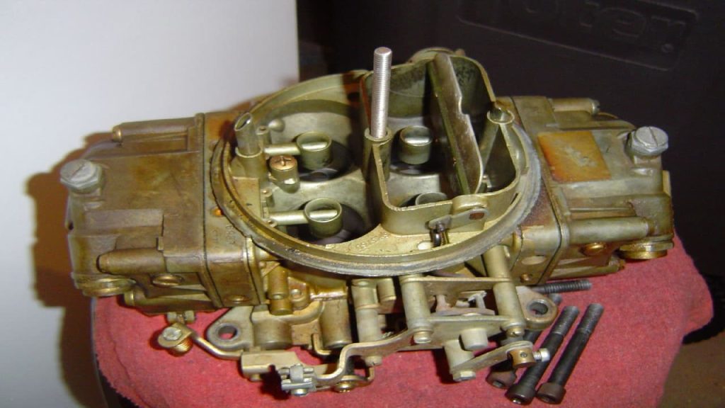 Holley Carburetor with brass-colored finish displayed on red foam padding, showing intake venturis and fuel adjustment mechanisms