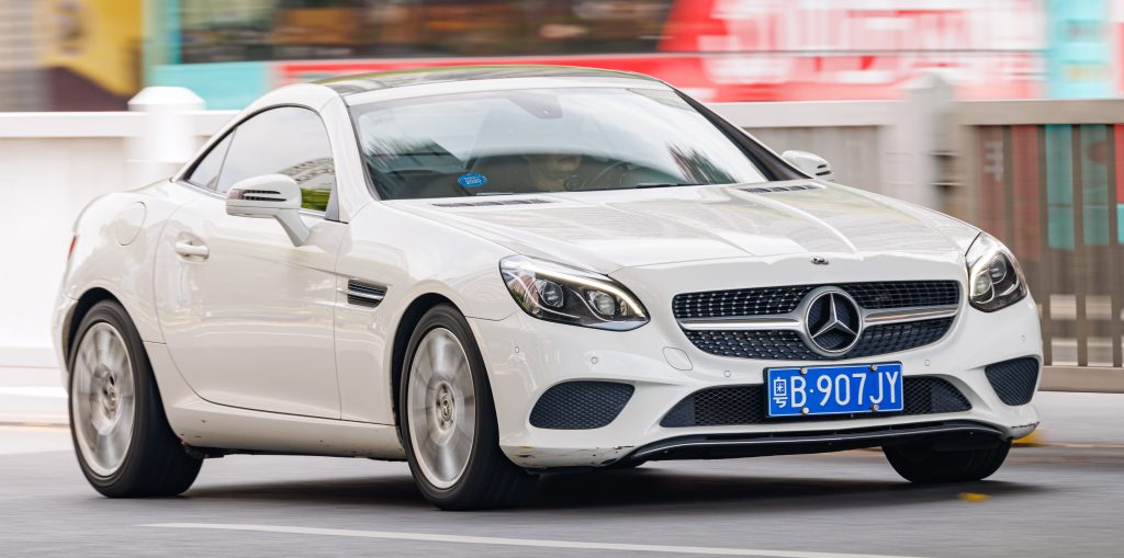 White Mercedes-Benz SLC convertible with blue Chinese license plate driving on city street with colorful buildings in background