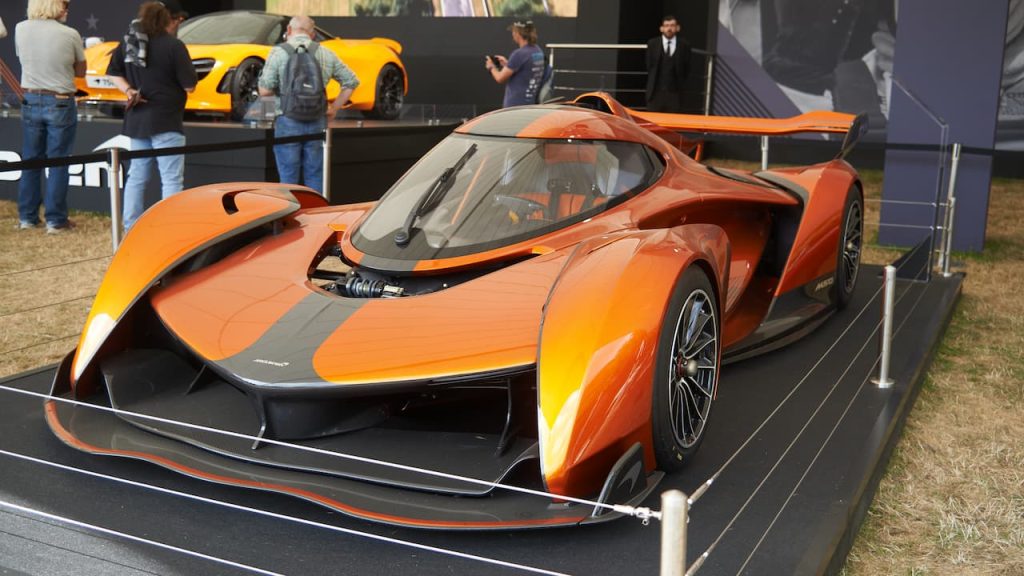 Orange McLaren Solus GT track-only hypercar with large rear wing displayed on platform at outdoor automotive show with spectators