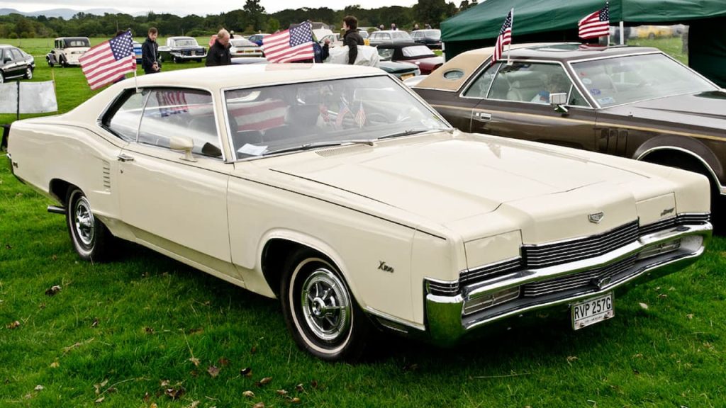 White vintage 1969 Mercury Marauder X-100 muscle car displayed at outdoor classic car show with American flags and spectators