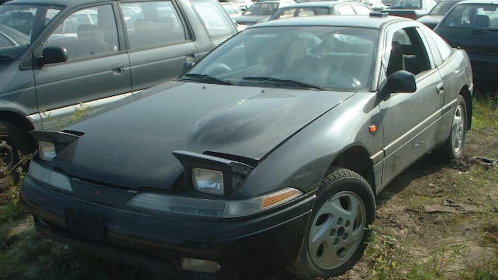 Silver late-model Mitsubishi Eclipse parked on cobblestone street in urban area with white SUV and buildings in background