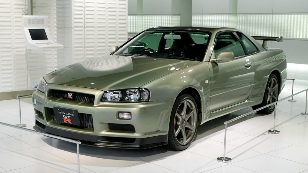 Green Nissan Skyline GT-R R34 on display in modern museum showroom with white walls and rope barriers