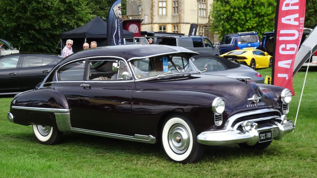 Vintage 1949 Oldsmobile Rocket 88 with whitewall tires displayed on grass at car show with Jaguar flag visible