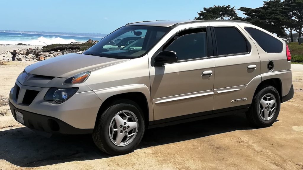 Beige Pontiac Aztek parked on beach with ocean waves, rocks, and cypress trees in background
