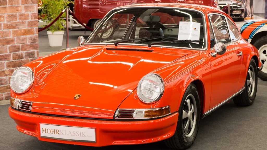Bright orange vintage Porsche 911 with "MOHR KLASSIK" dealer plate displayed in classic car showroom with brick walls