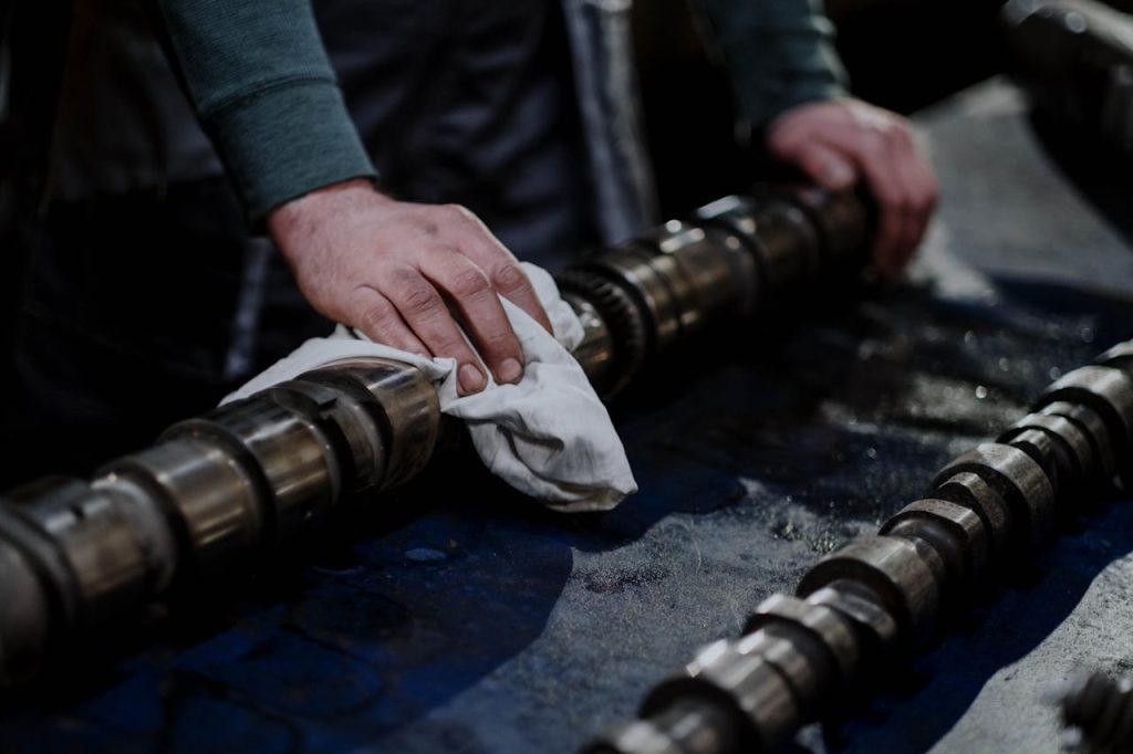 Mechanic's hands cleaning camshaft with white cloth, surrounded by multiple camshafts on blue work surface in automotive workshop