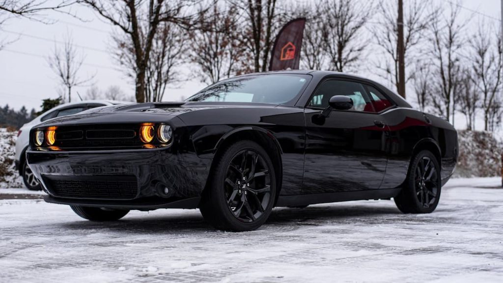 Dodge Challenger Black Ghost with illuminated yellow headlights parked on snowy surface against backdrop of bare winter trees