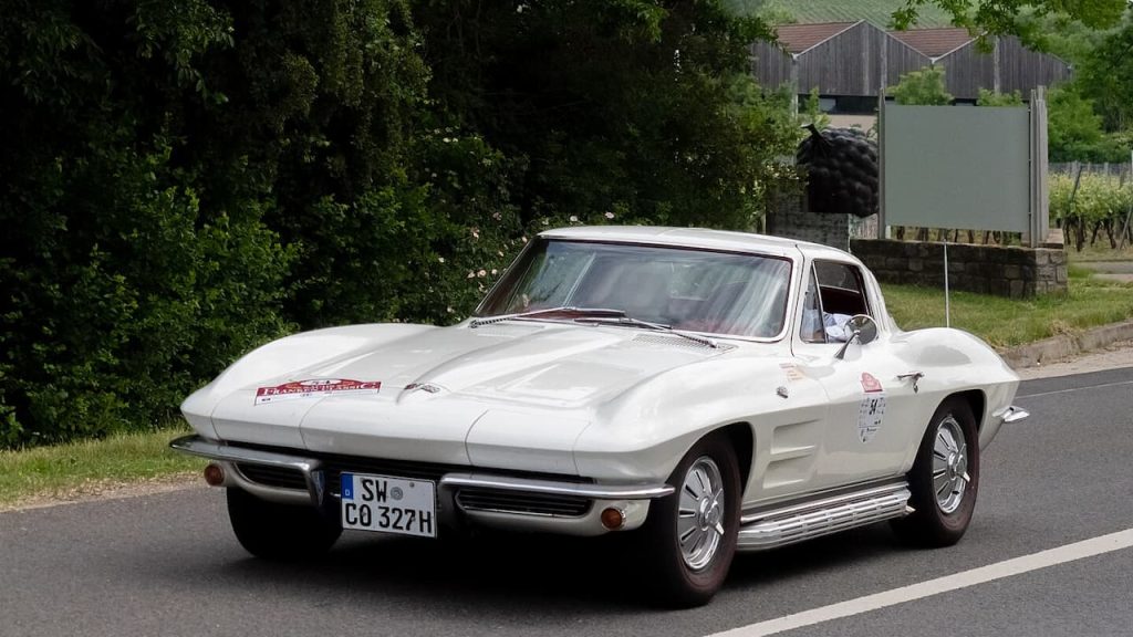 White Chevrolet Corvette C2 with split rear window and German registration SW-CO327H driving on rural road with green countryside