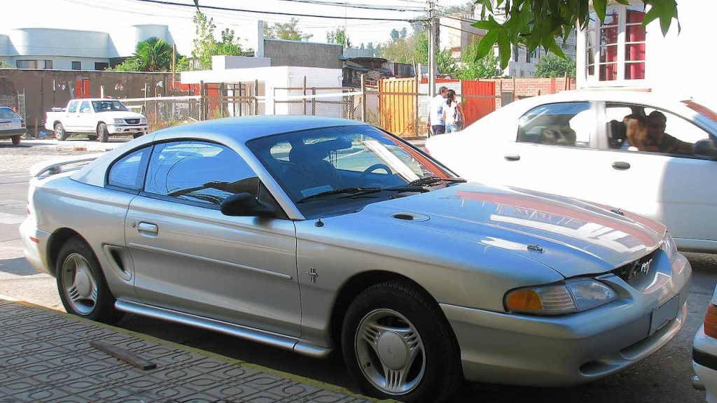 Silver Ford Mustang LX 1994 parked on cobblestone street in urban area with white SUV and buildings nearby