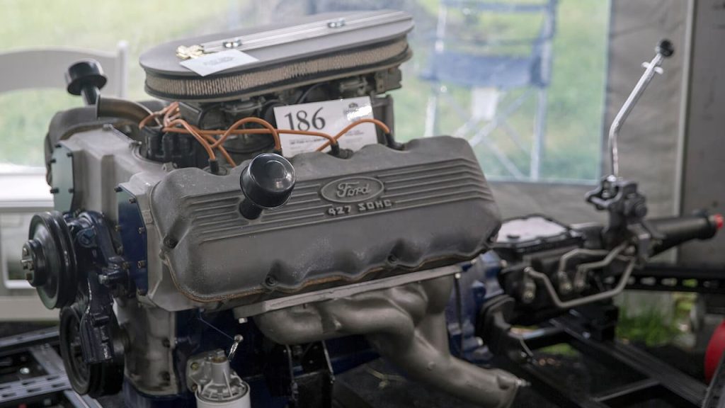 Ford 427 Cammer engine on display stand in workshop, showing valve cover, air cleaner, and exhaust headers with identification tag