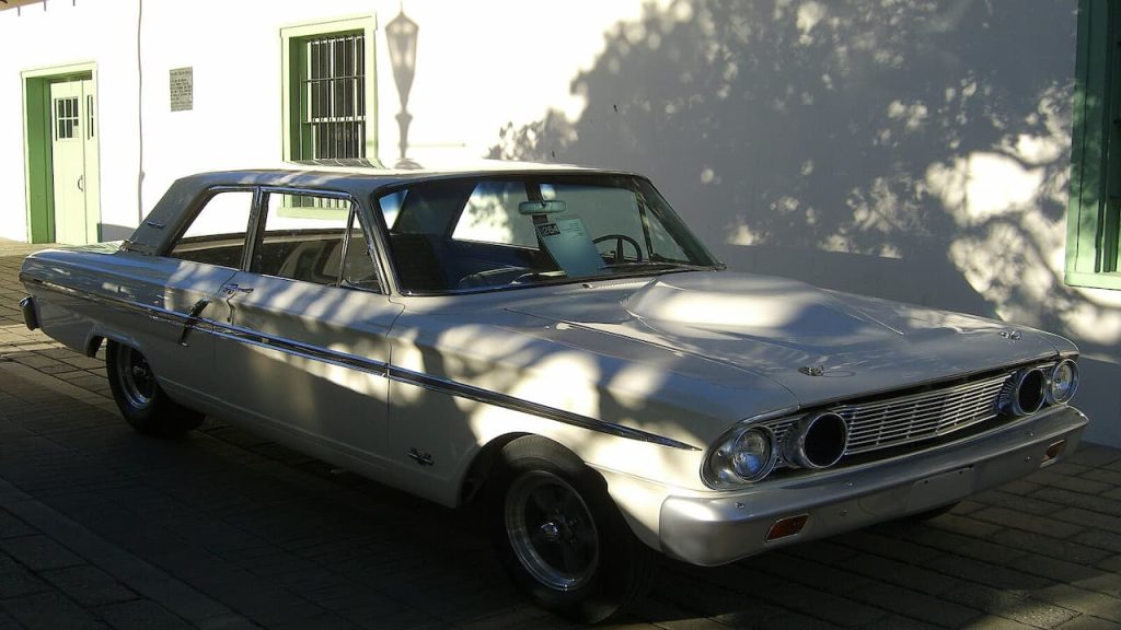 White 1964 Ford Thunderbolt muscle car parked on cobblestone street in front of white building with green trim